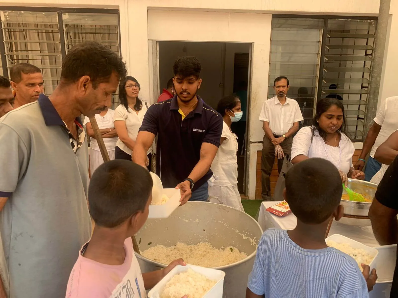 A person serving food from a large pot to a line of people, including children.